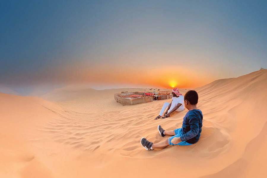 Two guests sitting on a desert dune at sunset